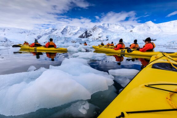 A group of kayakers in bright yellow kayaks are listening to a guide on a calm icy water surrounded by snow-covered mountains.