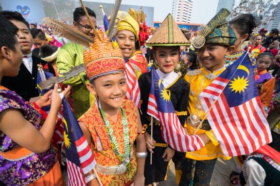 Children in traditional costumes, some with the Malaysian flag, pose at an outdoor event in Kuala Lumpur.