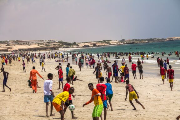 People enjoy the sandy beach in Mogadishu, Somalia, playing football and socializing under a clear sky.