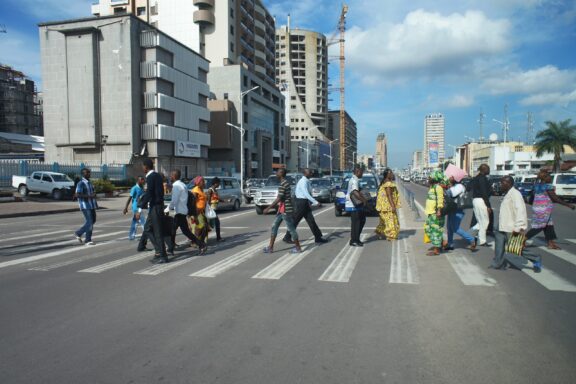 Pedestrians crossing a street on a zebra crossing in Kinshasa, Democratic Republic of Congo, with cars on the road and buildings in the background.