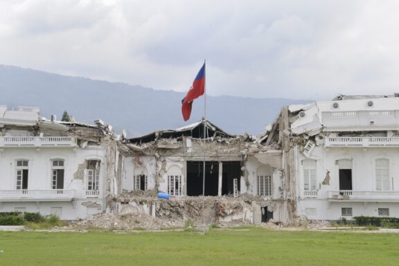 A damaged building with collapsed walls and debris in Port-au-Prince, Haiti, with a Haitian flag flying above.