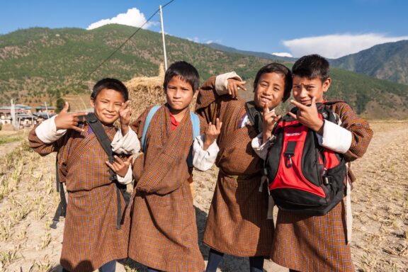 Four boys in traditional gho robes pose playfully in front of a mountainous backdrop in Punakha, Bhutan.