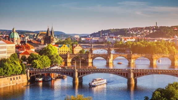 A picturesque spring sunset in Prague, highlighting the historic Charles Bridge, a cruising boat, and the city's glowing architecture.