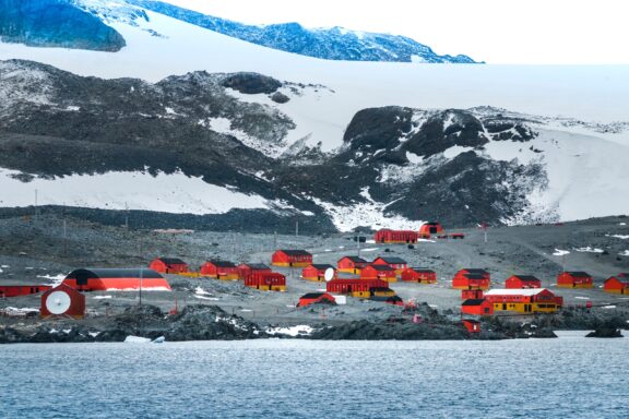 A research station with red buildings is situated along the rocky, snow-covered coastline of Antarctica.