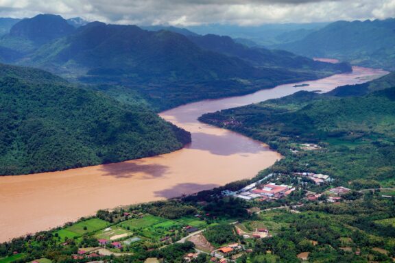 Soaring high above, the aerial view of the mighty Mekong River near Luang Prabang showcases the vastness and beauty of the waterway surrounded by lush green mountains and traditional Laos villages.