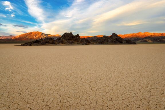 A sunset at Racetrack Playa in Death Valley National Park, showcasing a dry lake bed, sunlit mountains, and a cloudy sky.