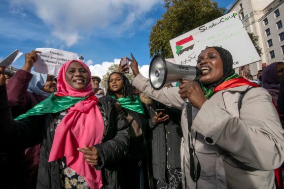 Sudanese political supporters, some in colorful headscarves, demonstrate under a clear sky with flags and banners in the background.