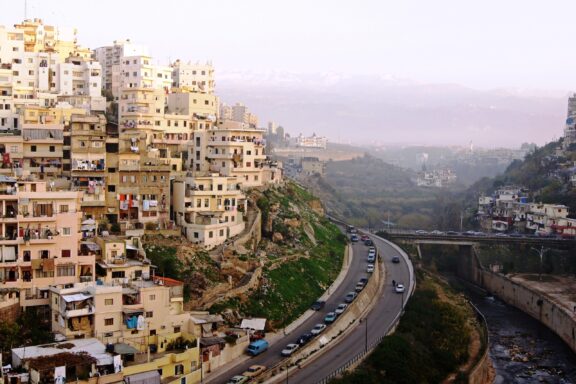 A winding road through a densely built hillside in Tripoli, Lebanon, with closely packed buildings and distant hazy mountains.