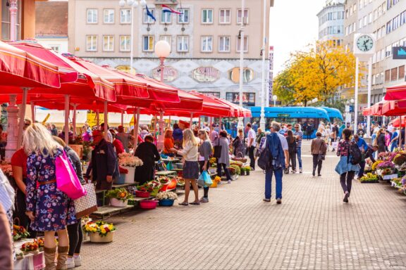 A bustling outdoor market in Zagreb, Croatia, with vendors under red canopies selling a variety of goods to people browsing the stalls.