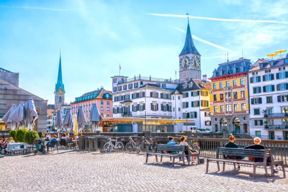 People enjoy a sunny day in Zurich, Switzerland, amidst colorful buildings and church spires.