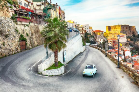 A small, blue car drives along a winding road in the small beachside town of Scilla, Italy.