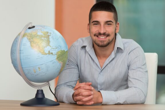 World globe on a desk, smiling man sitting next to the globe.