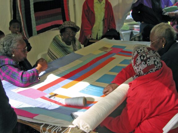 Women from Gee’s Bend in Wilcox County sit around a quilting frame.