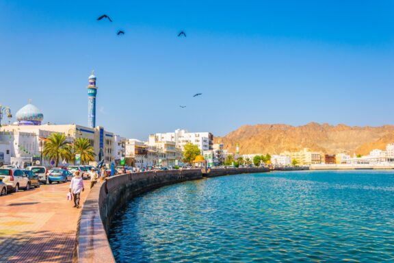 People walk along a promenade next to the water in Muscat, Oman on a sunny day.