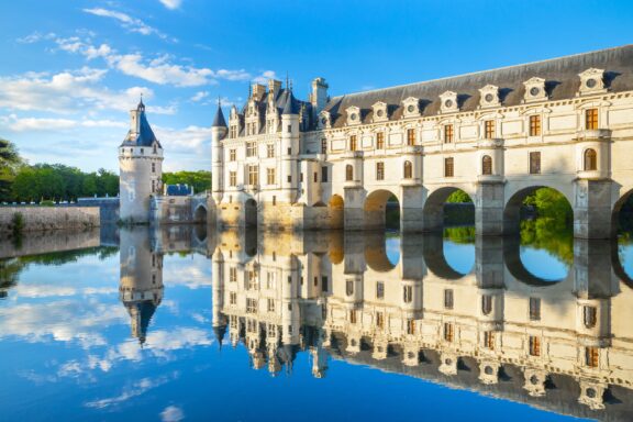 The Château de Chenonceau in France reflects in the water on a mostly clear day. 