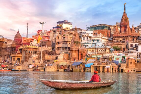 A man sits in a small boat on the Ganges River in Varanasi, India at sunset.