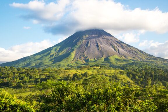 A cloud covers the top of Arenal Volcano, which towers over surrounding forests.