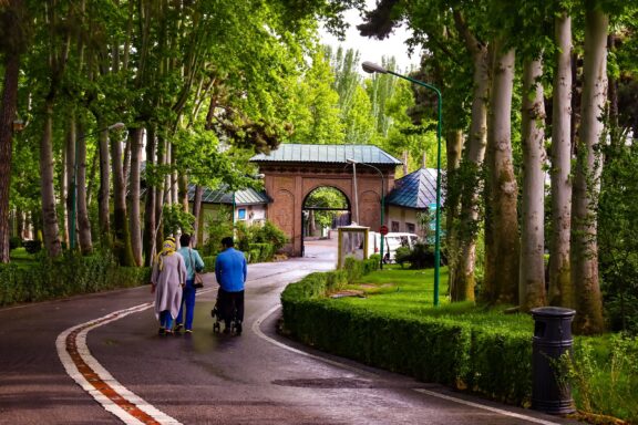 Three People Walking Towards a Gate Entrance Surrounded by Green Trees The grand entrance gate to the Sa'dabad Palace Complex
