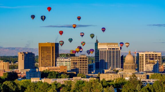 Colorful hot air balloons hover above the skyline of Boise, Idaho in Ada County at golden hour on a clear day. 