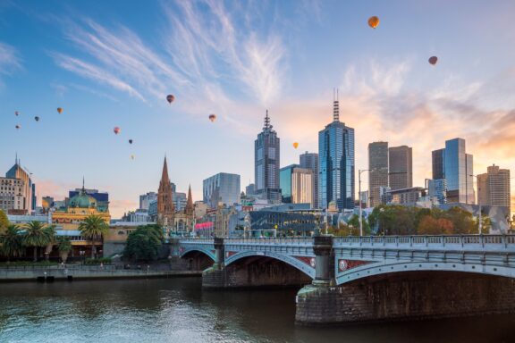 Hot air balloons hover over the Melbourne skyline near dusk.