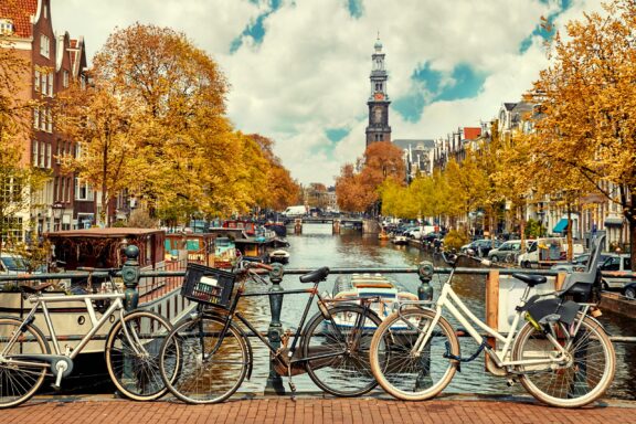 Bicycles lean against a canal bridge railing in Amsterdam, backed by trees with yellow leaves.