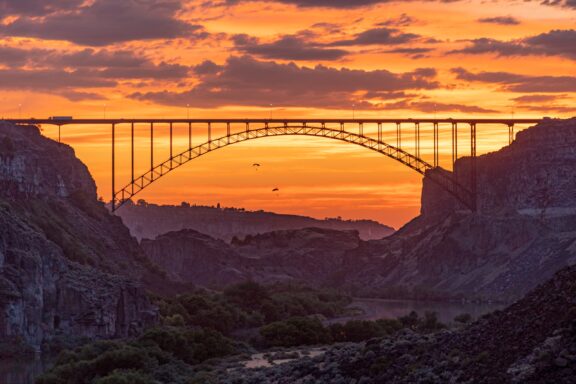 Two base jumpers parachute down from Perrine Bridge in Twin Falls, Idaho with a sunset sky in the background.