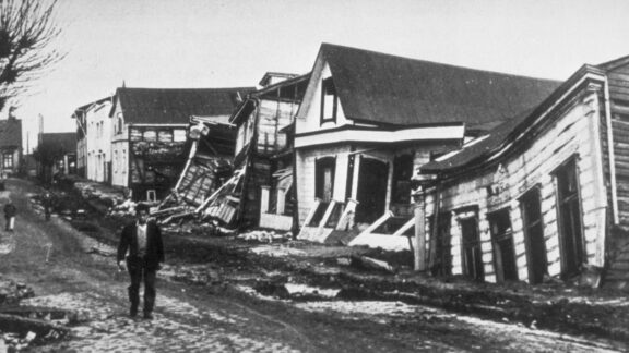 A man walks down a road lined with damaged wooden houses in a black-and-white photo of Valdivia, Chile in 1960.