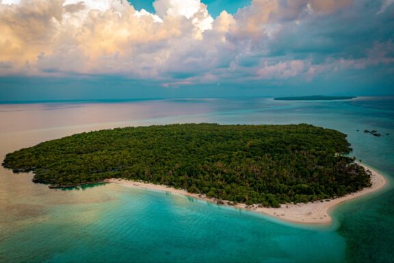 The small island of Anano in the Banda Sea is topped with trees and bordered by a small, sandy beach.