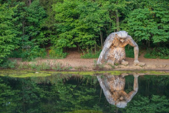 A view of a wooden sculptor sitting next to a lake from across the water in Bullitt County’s Bernheim Forest.