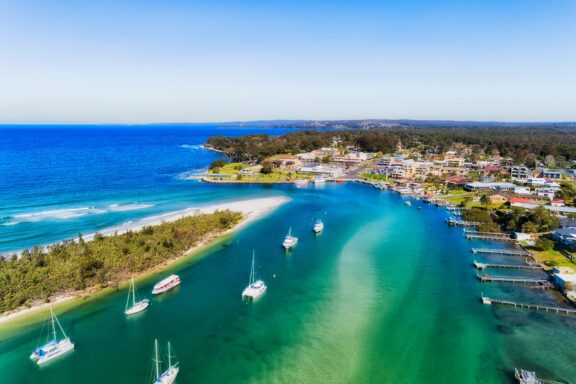 Aerial View of Currambene Creek Flowing into Jervis Bay with Moored Boats and Coastal Town An aerial view of sailboats floating in Curambene Creek near the ocean in Jervis Bary, Australia.