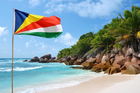 Seychelles flag waving on a clear day at Anse Cocos beach with turquoise water and granite boulders.