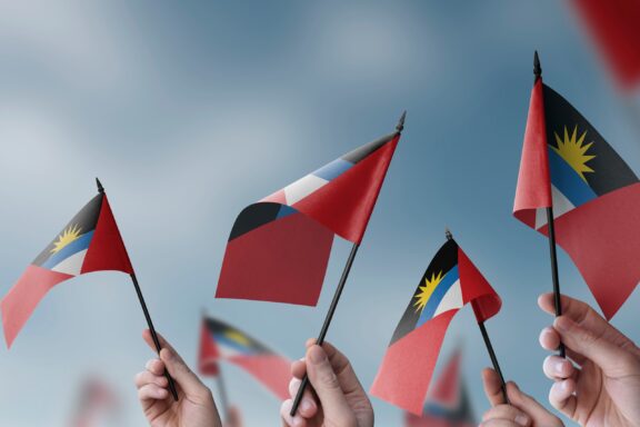 Multiple hands waving the flags of Antigua and Barbuda against a cloudy sky.