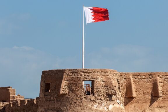 Flag of Bahrain waving above the ancient fort walls under a clear blue sky.