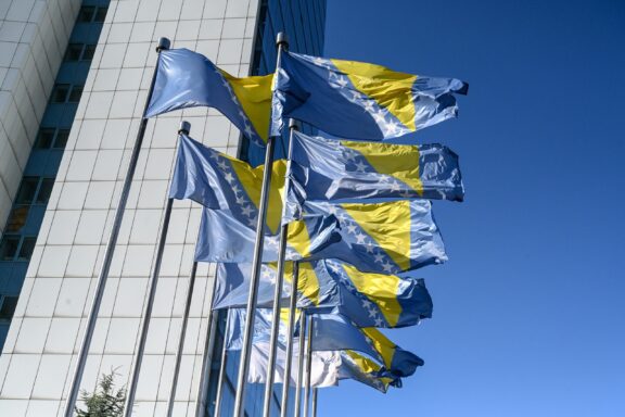 Bosnian flags fluttering in front of a government building under a clear blue sky.