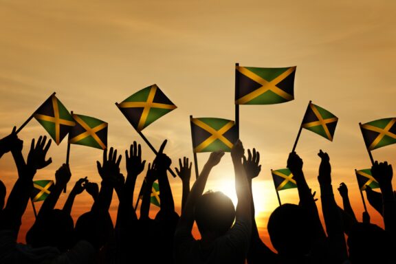 Multiple Jamaican flags waving against a sunset sky with silhouetted hands raised in the air.