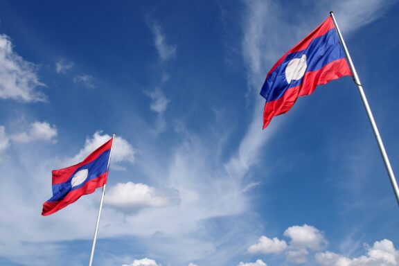 Two Laos flags waving against a blue sky with clouds.