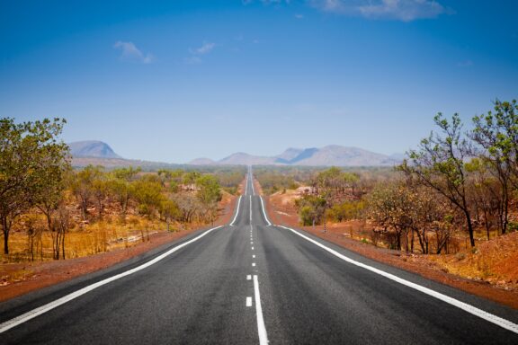 An empty road, bordered by red earth, stretches into the distance in Kimberly, Western Australia.