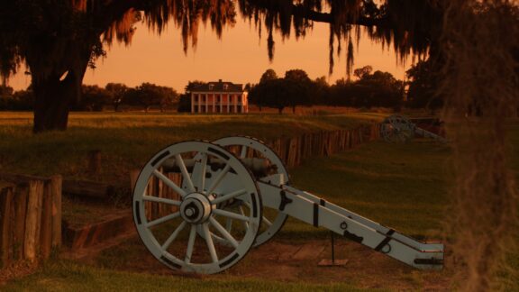 A canon sits in the foreground and the Beauregard Houe stands in the distance on the Chalmette Battlefield at dusk.