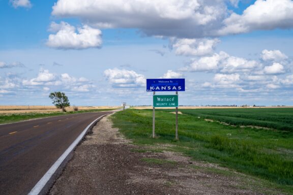 A “Welcome to Kansas” sign marks the Wallace County line along a highway surrounded by open fields.