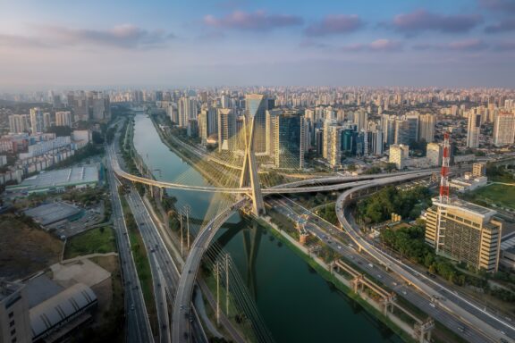 Aerial View of a Modern Cable-Stayed Bridge Spanning a River in a Bustling Cityscape at Dusk An aerial view of the Octavio Frias de Oliveira bridge and surrounding city in São Paulo, Brazil.