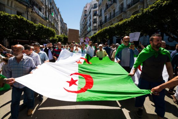 Protesters marching on a sunny day while carrying a large Algerian flag.