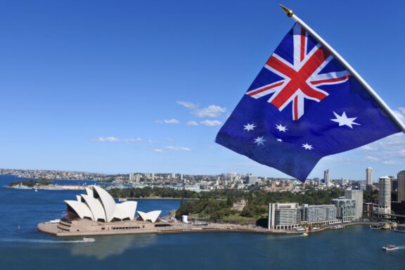 Australian flag waving in the foreground with the Sydney Opera House and city skyline in the background.