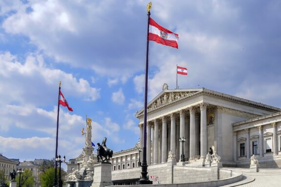 Austrian flags fluttering in front of the Parliament building under a cloudy sky.