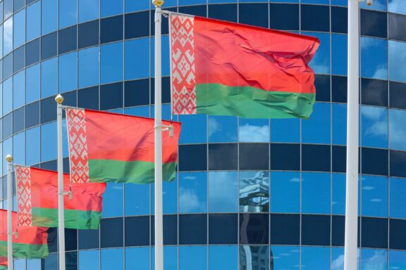 Belarusian flags fluttering in front of a glass building with a reflection of the sky.