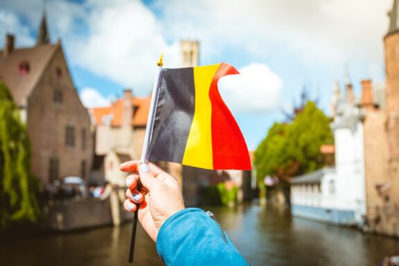 A person's hand holding a small Belgian flag with the Belfort tower and a canal in Bruges in the background.