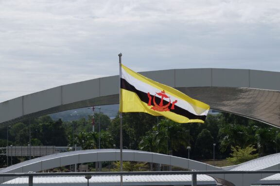 Brunei flag waving in front of a modern airport terminal under a cloudy sky.