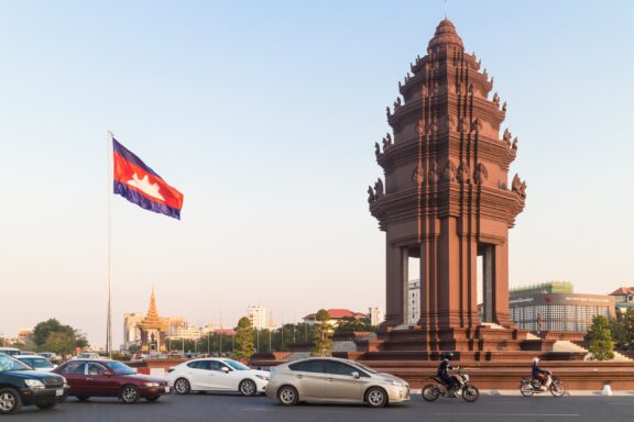 Cambodian Flag and Independence Monument in Phnom Penh Cambodian flag waving beside a traditional tower with vehicles parked in front on a clear day.