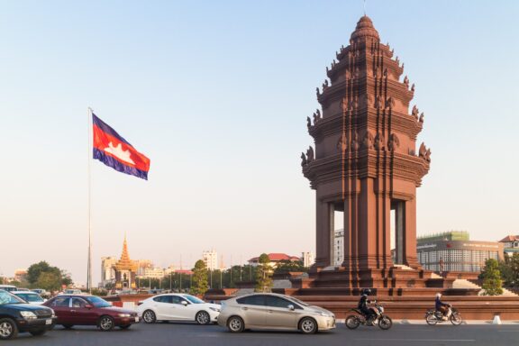 Cambodian flag waving beside a traditional tower with vehicles parked in front on a clear day.