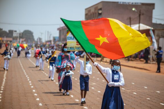 Children in uniform marching in a parade with one child carrying the Cameroon flag.