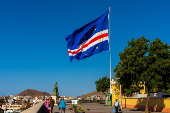 Cape Verde flag waving against a clear blue sky with people walking in the foreground and a cityscape in the background.